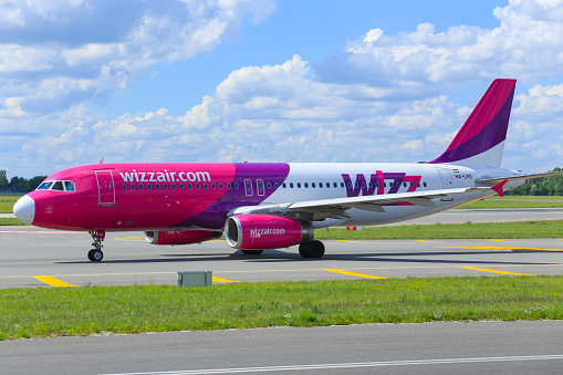 Sunny day, A Wizz Air Airbus A320 aircraft is taking off from the runway of the Lech Walesa Airport in Gdansk, Poland. The aircraft is white with a pink and purple tail and “Wizz Air” written in pink on the side. The aircraft is lifting off the ground with its nose up and its landing gear retracting. The background consists of a blue sky and a few buildings and trees in the distance.