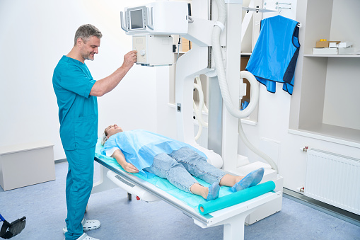 Friendly doctor in a blue suit prepares a woman for an x-ray of the pelvis on a modern machine
