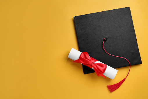 Graduation hat and diploma on yellow background, flat lay. Space for text