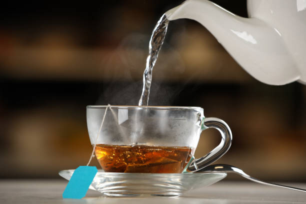 istock Pouring hot water into glass cup with tea bag on table against blurred background 1414764861