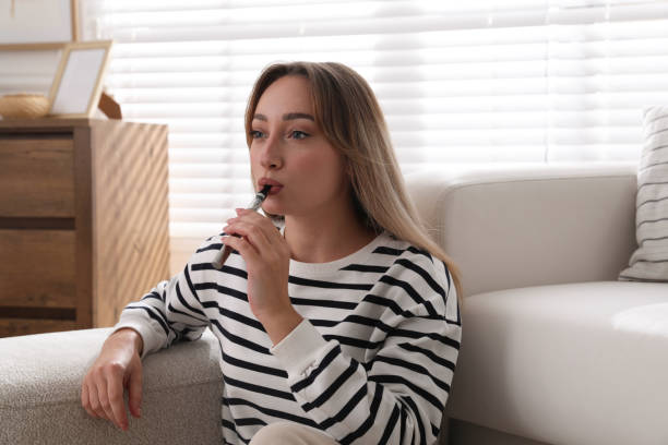 istock Beautiful young woman using electronic cigarette at home 1414765078