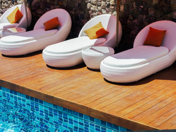 istock Poolside decoration with empty modern white sunbeds with orange pillows on wood plank deck terrace on stone wall background near the swimming pool. Empty outdoor pool on sunshine day in summer. 1414767303