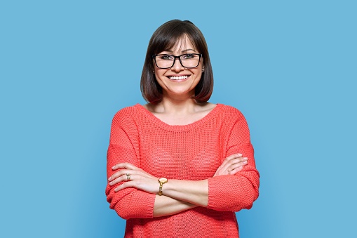Attractive middle aged woman looking at camera on blue color background. Smiling confident 40s female with glasses, crossed arms in red casual clothes. Lifestyle, confidence, calmness, mature people