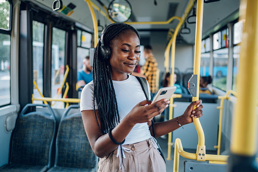 Young African american woman wearing earphones and listening to music on a smartphone while standing alone on a bus. Traveling to work and enjoying a bus ride. Portrait of a beautiful black woman.