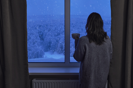 A woman stands at a winter window with trees in the snow. A woman with a cup of coffee looks out the evening window with a snowfall behind the glass