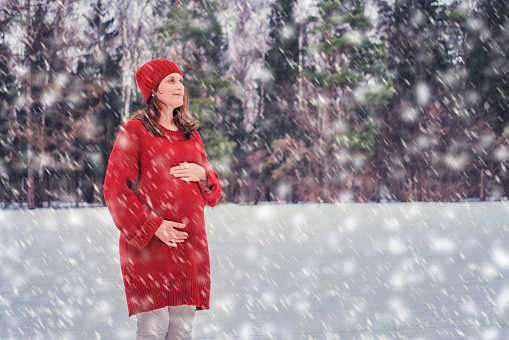 A happy pregnant woman in a knitted sweater and a hat walks in the park, a winter forest with trees in the snow