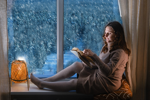 Young woman reading a book by the window in the light of the evening lamp. Woman sitting on the windowsill by the window with snow-covered trees in the winter forest