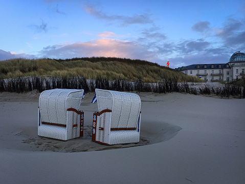 Two beach chairs on the beach of the island of Juist stand in the blown sand in the evening light. The sun illuminates the few clouds in the blue sky. The view goes to the dunes and partly it is free on the beach hotel Juist.
