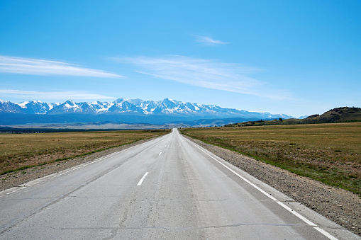 A straight road goes into the distance into a snowy mountain range. Panoramic landscape background. Altai mountains