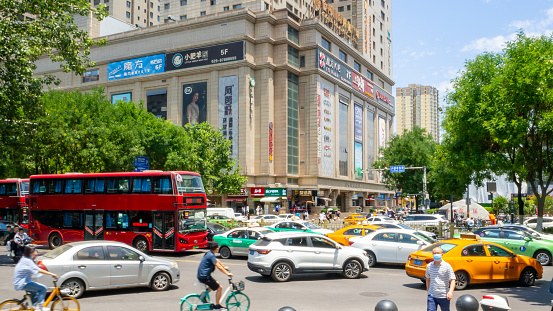 Xi'an, China - July 20, 2022: People riding bicycles among the city traffic. There is a commercial building with business signs in the background.