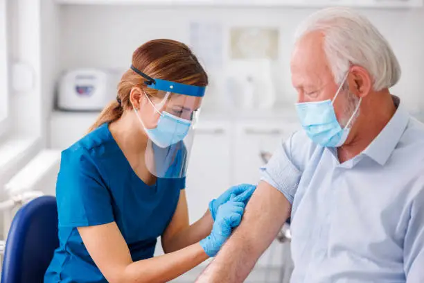 Elderly man wearing protective medical face mask receiving covid 19 or seassonal flu vaccine at medical clinic; doctor or nurse touching and disinfecting the vaccine application spot
