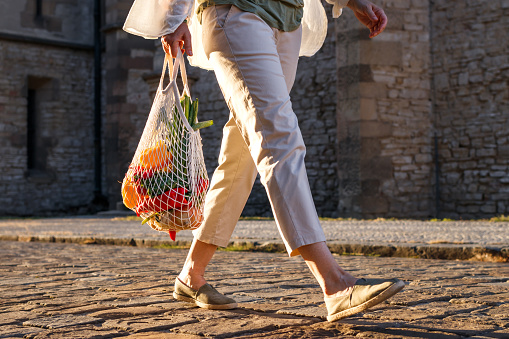 Woman walks on street and carrying reusable mesh bag with vegetable after shopping in motion. City life