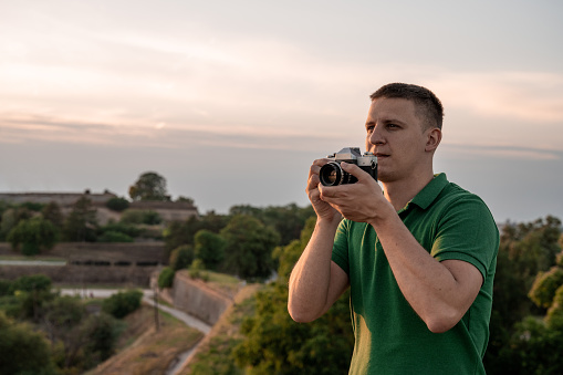 Handsome young man enjoying the view. He is holding old retro camera. Fortress and sunset in background.