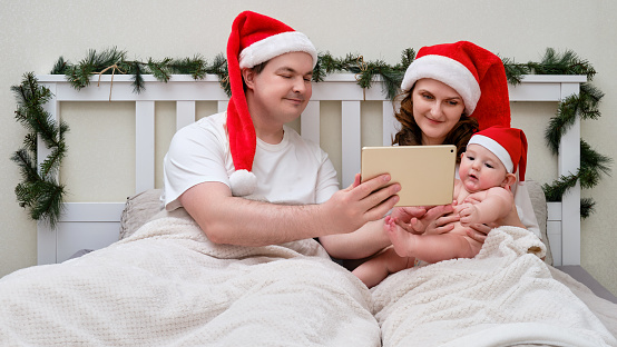 Happy man and woman with child take photo on tablet in bed decorated for christmas and new year, family couple in home bedroom