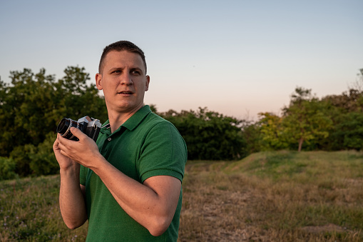 Man looking aside while holding old-fashioned camera in arms. Nature in background