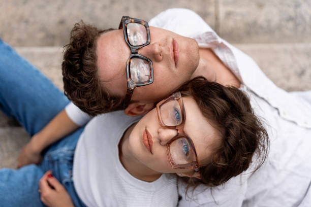 Two attractive students man and woman wearing stylish glasses sitting floor Portrait of two attractive students man and woman wearing stylish glasses sitting floor outdoor and smiling, head to head cheek to cheek, break relax between lesson, work, projects Eyeglasses concept cheek to cheek stock pictures, royalty-free photos & images