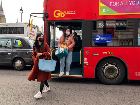 Two women, one of whom is carrying a small pet dog, getting off a red double-decker London bus in Westminster, London. Both women are of Asian ethnicity and are wearing face masks.