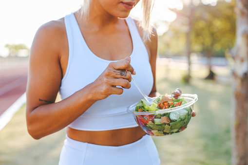 Young female athlete eating healthy after training. Athlete exercising outdoors. Lifestyle health and wellness. Outdoor public park, natural sunlight.
