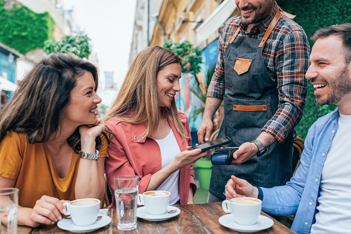 Young woman paying contactless using smartphone in sidewalk cafe, with friends. Customer paying through mobile phone using contactless technology