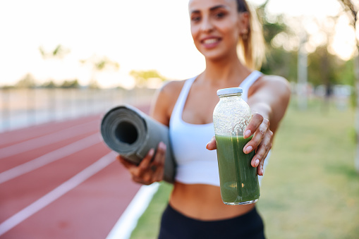 Sporty woman drinking detox green smoothie in the Park. Green smoothie woman drinking plastic cup breakfast meal takeaway to go after morning run on city streets. Healthy lifestyle sporty person pov of hand holding glass with running shoes feet selfie.