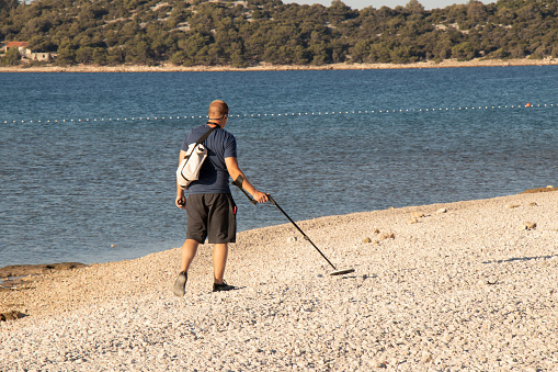 Vodice, Croatia - July 20, 2022: Person walking on the empty pebble beach using outdoor metal detector device
