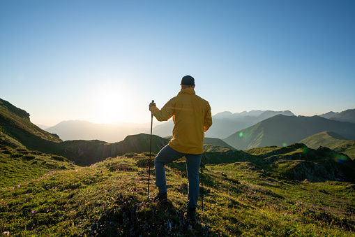 Rear view of the hiker stands alone with hiking poles in the mountains at sunset