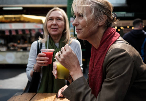 retrato de dos amigas maduras sentadas en una mesa en un mercado callejero bebiendo jugos. - drinking straw drinking juice women fotografías e imágenes de stock