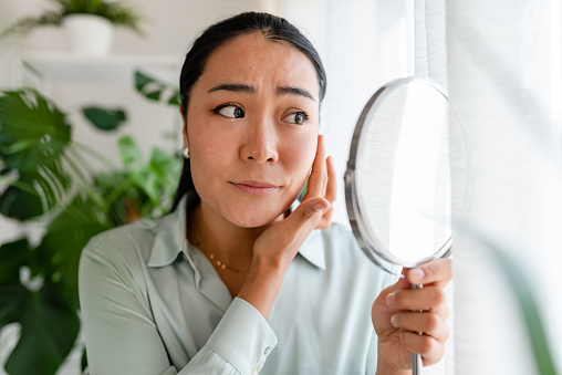 Young Asian woman looking herself in the mirror. Worried about acne caused by wearing a mask. Maskne. Problems with acne.