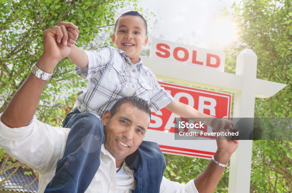 Happy Young Hispanic Father and Son In Front of Sold For Sale Real Estate Sign. Selling Stock Photo