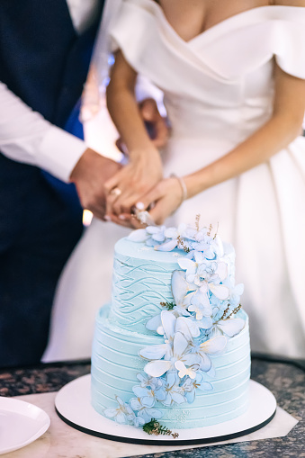 The bride and groom are holding a knife and cutting a stylish wedding cake with blue butterflies decorations at a restaurant reception.