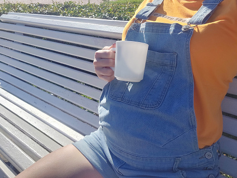 Pregnant young woman sitting on a bench and drinking coffee from white cup . Unhealthy bad food and drink during pregnancy. Inscription is cautiously hot.