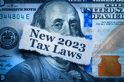 A torn piece of paper telling of the new 2023 tax laws rests on top of a one hundred dollar bill.