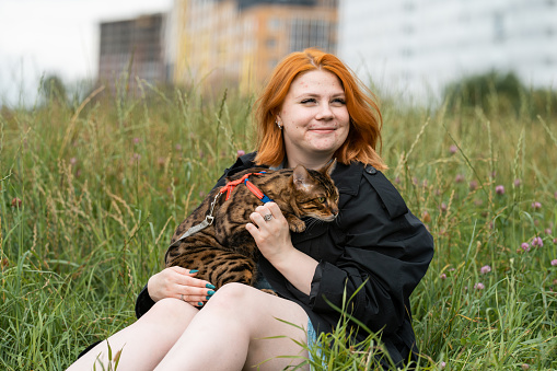 Young red head woman with acne sitting in grass with her domestic tabby cat with leash.