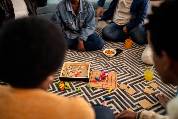 Cropped view of an African-American family playing chinese checkers. The game board is in the middle on a coffee table. The faces of two teenage sisters smiling at each other are out of focus in the foreground. The focus is on the board and dad's hand.