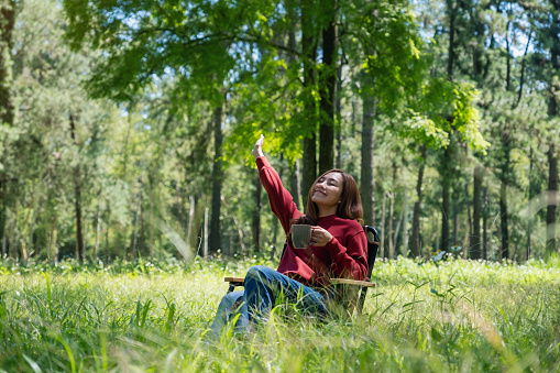 Portrait image of a happy young asian woman raising hand while drinking coffee and sitting on a chair in the park