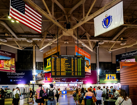 Boston, Massachusetts, USA - August 11, 2022: South Station, officially The Governor Michael S. Dukakis Transportation Center at South Station, is the largest railroad station and intercity bus terminal in Greater Boston. Located at the intersection of Atlantic Avenue and Summer Street in Dewey Square, Boston, the historic station building was constructed in 1899. View of the main concourse with train arrival and departure information display in the center and American and Massachusetts State flags hanging from the ceiling.