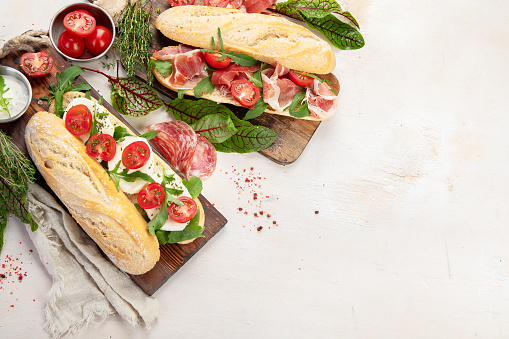 Two baguette sandwiches with salami, mozzarella cheese, lettuce, tomatoes and ham on a cutting boards. Long subway sandwiches on a white background. Top view with copy space.