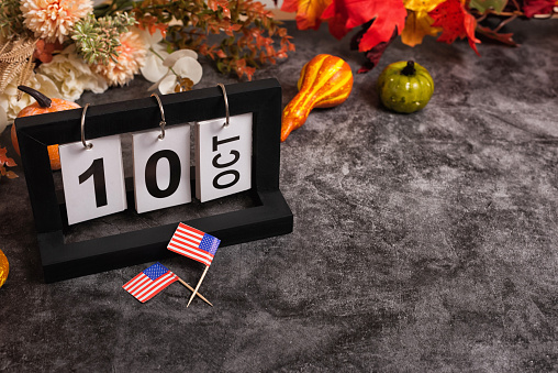Calendar with October 10th 2022, Columbus Day. Autumn festival composition and background with stars and strips flags. Gray concrete table decorated with pumpkins fall flowers and leaves