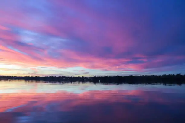 Hot pink and baby blue cotton candy cloudscape reflecting across the water. Tranquil calm water surface to the treelined horizon.