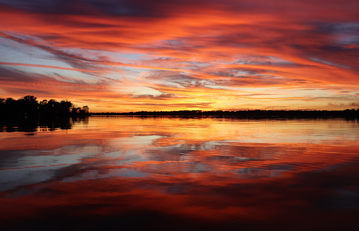 Sunset on Scott's Point at Lake Houston, Texas, Red swirl cloudscape reflect across lake to the horizon. Treelined point juts out into view on calm waters.