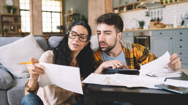 istock Couple calculating bills at home using calculator.  Couple checking bills and documents while calculating finances sitting in the living room. Man and woman at home analyzing their finance with documents. 1414921454
