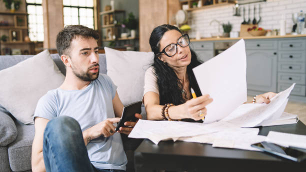 istock Couple calculating bills at home using calculator.  Couple checking bills and documents while calculating finances sitting in the living room. Man and woman at home analyzing their finance with documents. 1414921455
