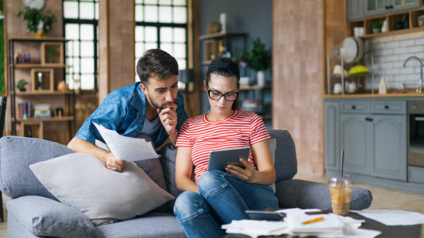 coppia che calcola le bollette a casa usando tablet e calcolatrice. giovane coppia che lavora al computer mentre calcola le finanze seduta sul divano. giovane con moglie a casa che analizza le loro finanze con documenti. - women document looking couple foto e immagini stock