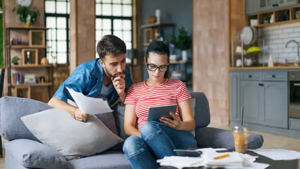 Photo of Couple calculating bills at home using tablet and calculator. Young couple working on computer while calculating finances sitting on couch. Young  man with  wife at home analyzing their finance with documents.