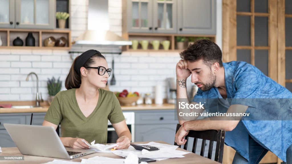 Frustrated concerned young couple calculating overspend budget, doing paperwork job at laptop, talking about financial problems, insurance, mortgage, fees, loan conditions, bankruptcy, economic inflation Couple - Relationship Stock Photo