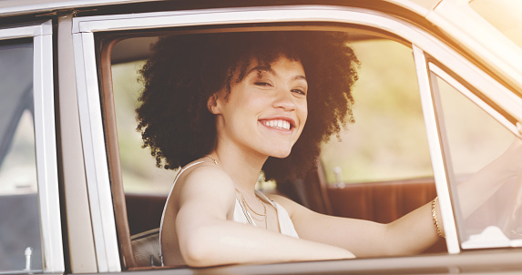Retro and trendy woman driving car on a road trip with vibrant and smiling expression on her face looking carefree and cool in summer. Afro American girl with cool vintage style taking a drive alone