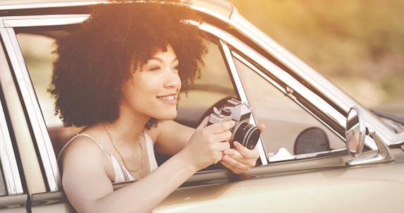 Beautiful, attractive woman on a holiday trip in the car outdoors during sunset. Happy, smiling and relaxed female on a vacation journey, holding a camera and taking a moment to admire the view.
