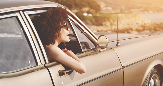 A carefree young woman on a road trip feeling relaxed, free and happy while putting her head out a vintage window. A young, smiling and afro hair female enjoying freedom while driving on a summer day