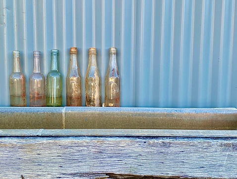 Outdoor country still life of empty antique vase and drinking glass bottles lined up against corrugated iron shed above metal livestock drinking trough with rustic wood frame in country Australia