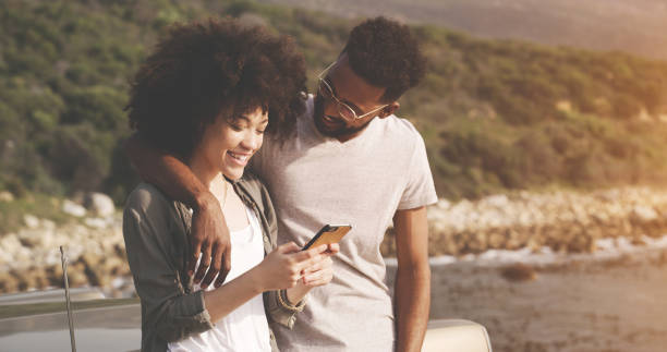 istock Carefree black couple bonding, making a booking online on a phone, talking and affectionate. Travelling boyfriend and girlfriend enjoying time together, taking a break from road trip to enjoy view 1414951270